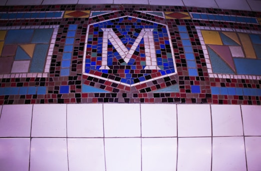 A colorful mosaic featuring an arrangement of small, square tiles forming an abstract design. At the center, the letter 'M' stands out prominently, surrounded by geometric patterns in various hues. The background consists of larger square tiles.