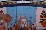 A colorful mural depicting a lively street scene with people walking towards a large building. Several birds fly in the sky above. The mural is set against a blue sky and is bordered by vibrant geometric patterns and floral decorations.