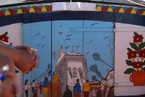 A colorful mural depicting a lively street scene with people walking towards a large building. Several birds fly in the sky above. The mural is set against a blue sky and is bordered by vibrant geometric patterns and floral decorations.