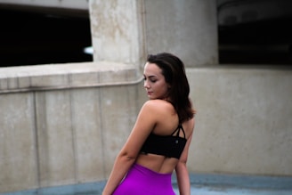 A woman stands confidently in an outdoor setting, wearing a black sports bra and vibrant purple leggings. The background consists of concrete structures with a modern, minimalist design.
