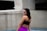A woman stands confidently in an outdoor setting, wearing a black sports bra and vibrant purple leggings. The background consists of concrete structures with a modern, minimalist design.