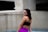A woman stands confidently in an outdoor setting, wearing a black sports bra and vibrant purple leggings. The background consists of concrete structures with a modern, minimalist design.