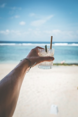 A hand holding a glass of le coquers beverage with the sea and horizon blurred in the background.