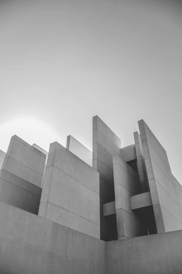 A striking black and white photograph of a modern, geometric building with sharp angles and intersecting planes. The structure has a minimalist design with a series of rectangular solid shapes positioned at varying heights, contributing to an abstract and sculptural aesthetic.