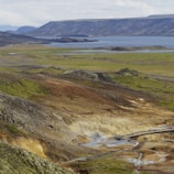 A vast landscape featuring a geothermal area with steam rising from the ground, surrounded by green and brown hills. A pathway crosses the geothermal area, where several people are walking and observing the environment. In the background, a serene body of water extends toward distant mountains under a cloudy sky.