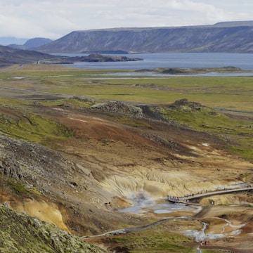 A vast landscape featuring a geothermal area with steam rising from the ground, surrounded by green and brown hills. A pathway crosses the geothermal area, where several people are walking and observing the environment. In the background, a serene body of water extends toward distant mountains under a cloudy sky.