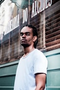 A man with braided hair and a nose piercing stands in front of a barbershop with a serious expression. He is wearing a white t-shirt, and the barbershop is named 'Barbearia do Alcides' with its name visible on the window behind.