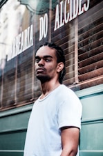 A man with braided hair and a nose piercing stands in front of a barbershop with a serious expression. He is wearing a white t-shirt, and the barbershop is named 'Barbearia do Alcides' with its name visible on the window behind.