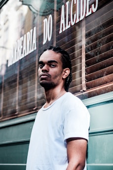 A man with braided hair and a nose piercing stands in front of a barbershop with a serious expression. He is wearing a white t-shirt, and the barbershop is named 'Barbearia do Alcides' with its name visible on the window behind.