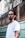 A man with braided hair and a nose piercing stands in front of a barbershop with a serious expression. He is wearing a white t-shirt, and the barbershop is named 'Barbearia do Alcides' with its name visible on the window behind.