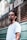 A man with braided hair and a nose piercing stands in front of a barbershop with a serious expression. He is wearing a white t-shirt, and the barbershop is named 'Barbearia do Alcides' with its name visible on the window behind.
