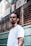 A man with braided hair and a nose piercing stands in front of a barbershop with a serious expression. He is wearing a white t-shirt, and the barbershop is named 'Barbearia do Alcides' with its name visible on the window behind.