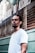 A man with braided hair and a nose piercing stands in front of a barbershop with a serious expression. He is wearing a white t-shirt, and the barbershop is named 'Barbearia do Alcides' with its name visible on the window behind.