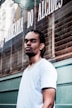 A man with braided hair and a nose piercing stands in front of a barbershop with a serious expression. He is wearing a white t-shirt, and the barbershop is named 'Barbearia do Alcides' with its name visible on the window behind.