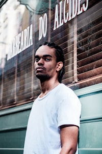 A man with braided hair and a nose piercing stands in front of a barbershop with a serious expression. He is wearing a white t-shirt, and the barbershop is named 'Barbearia do Alcides' with its name visible on the window behind.