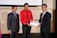 Three men in formal attire are standing indoors. The man in the center is wearing a red shirt and is receiving a certificate from the man on the right, who is dressed in a dark suit. The man on the left is also wearing a suit and is smiling towards the camera. They appear to be attending a professional event or ceremony.