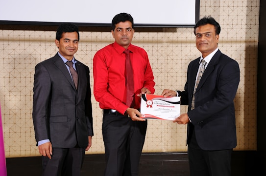 Three men in formal attire are standing indoors. The man in the center is wearing a red shirt and is receiving a certificate from the man on the right, who is dressed in a dark suit. The man on the left is also wearing a suit and is smiling towards the camera. They appear to be attending a professional event or ceremony.