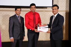 Three men in formal attire are standing indoors. The man in the center is wearing a red shirt and is receiving a certificate from the man on the right, who is dressed in a dark suit. The man on the left is also wearing a suit and is smiling towards the camera. They appear to be attending a professional event or ceremony.