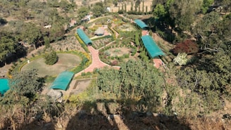An aerial view of a well-maintained garden with pathways lined by greenery and trees, and covered walkways with blue-green roofing. The garden has circular and rectangular lawn sections with manicured bushes and a variety of plants.