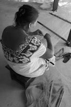 A person is sitting on a small stool, weaving on a traditional loom. The person's clothing features intricate patterns. Loose fibers and weaving tools are placed on the ground near their feet, which are barefoot. The setting appears simple, with a rough floor surface and minimal additional objects visible.