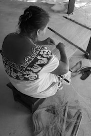 A person is sitting on a small stool, weaving on a traditional loom. The person's clothing features intricate patterns. Loose fibers and weaving tools are placed on the ground near their feet, which are barefoot. The setting appears simple, with a rough floor surface and minimal additional objects visible.