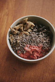 A bowl filled with various ingredients, including a pink smoothie-like base topped with chopped nuts or seeds, brown dried fruits, and possibly cacao nibs.