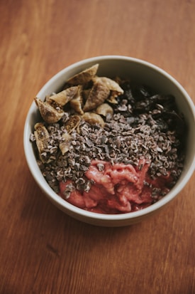 A bowl filled with various ingredients, including a pink smoothie-like base topped with chopped nuts or seeds, brown dried fruits, and possibly cacao nibs.