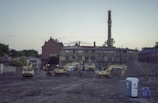 An urban construction site with several excavators positioned in front of a large brick building with an industrial chimney. The background includes other older structures, and the scene is set during twilight, with a dimly lit sky.