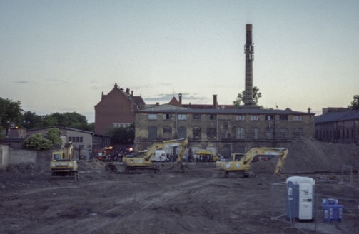 An urban construction site with several excavators positioned in front of a large brick building with an industrial chimney. The background includes other older structures, and the scene is set during twilight, with a dimly lit sky.
