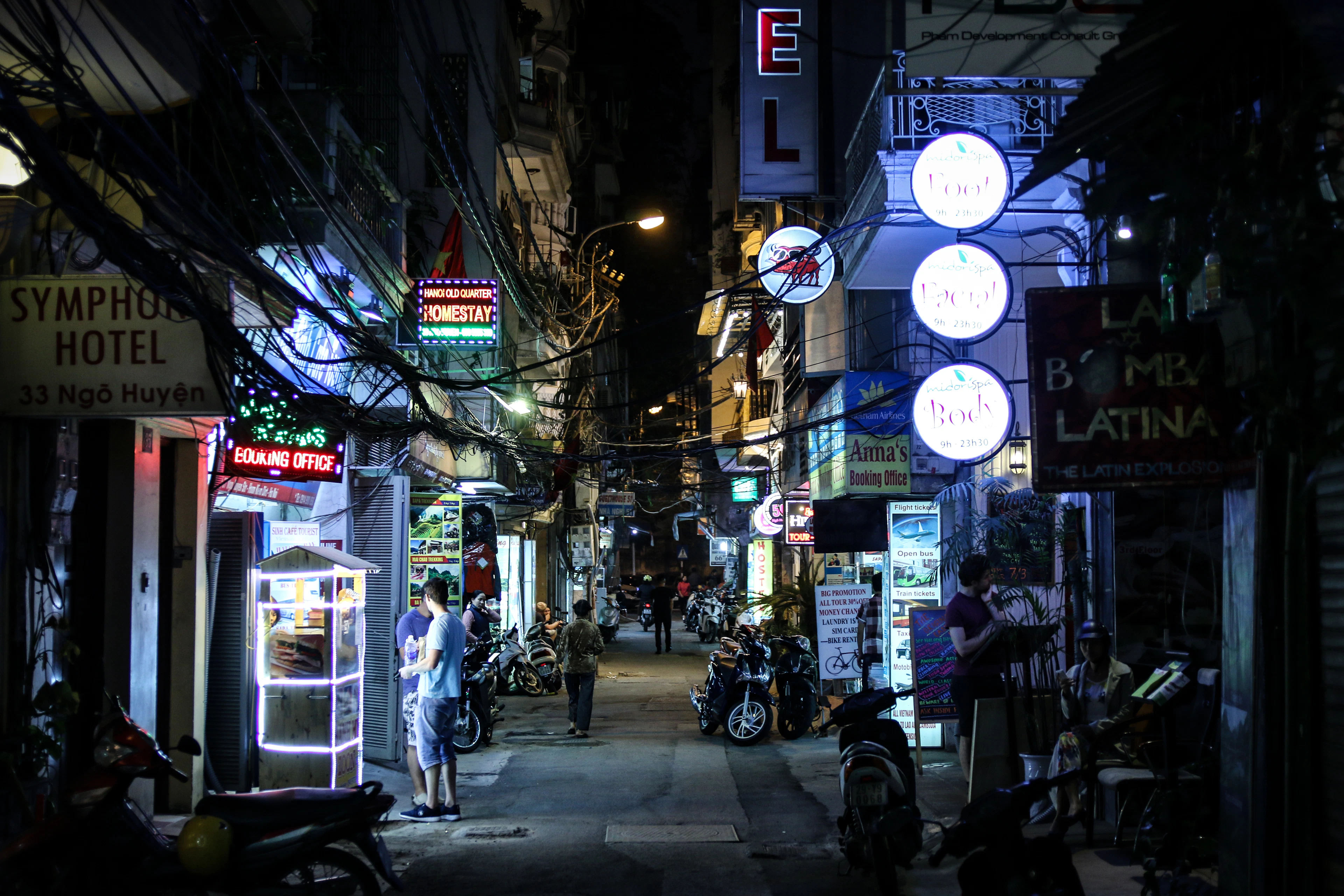 Neon shopfronts and motorbikes glow along a busy Hanoi Old Quarter street at night.