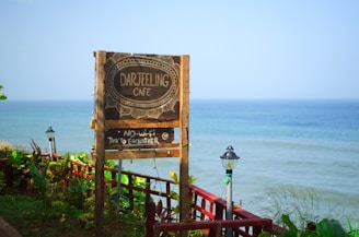 A rustic wooden signboard for 'Darjeeling Cafe' near a body of water with a 'No Wi-Fi, Talk to Each Other' message. The background features a serene ocean under a clear blue sky, with some greenery and lamp posts visible along a railing.