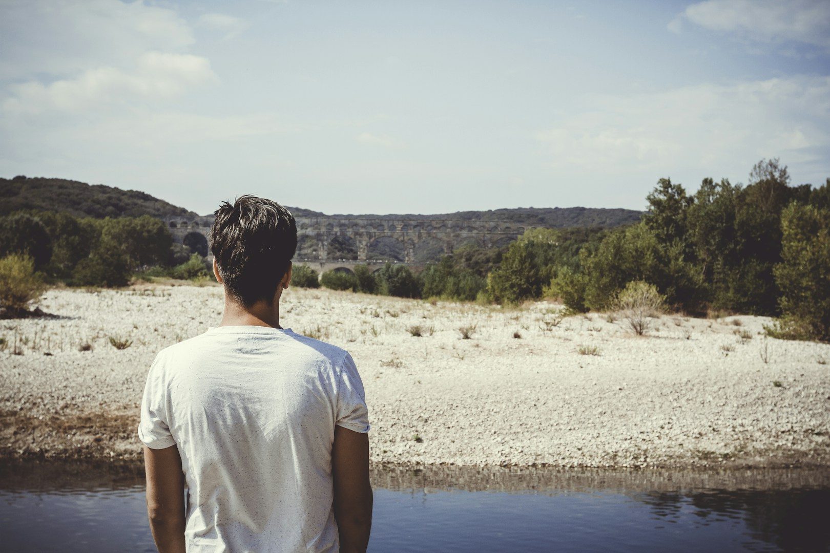 man standing at the edge of water, looking at a bridge in the distance