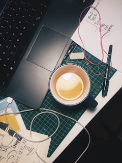A workspace featuring a laptop with a visible touchpad, a cutting mat, and a cup with a beverage, possibly tea or coffee. Artistic sketches and handwritten notes scattered, alongside tangled earphones, a pen, and other stationery items contributing to a creative and slightly cluttered atmosphere.