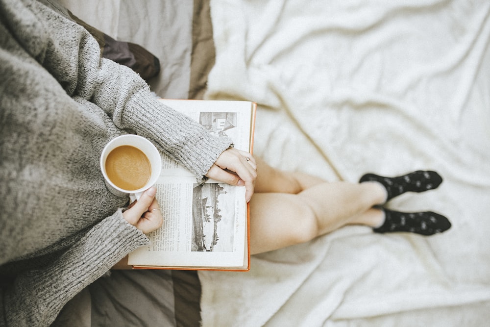 오른손에 커피 한 잔을 들고 무릎에 앉아 책을 읽고 있는 여성은 조명이 밝은 방에서 왼손으로 책을 펴고 있습니다