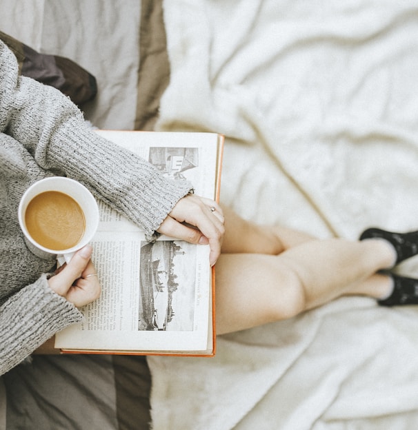 woman holding a cup of coffee at right hand and reading book on her lap while holding it open with her left hand in a well-lit room