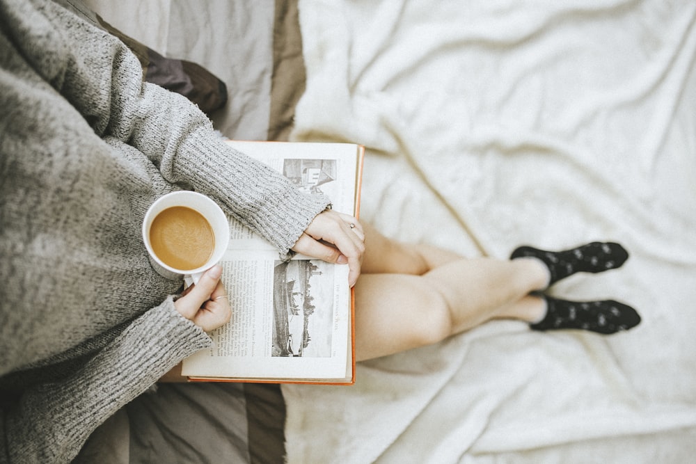 woman holding a cup of coffee at right hand and reading book on her lap while holding it open with her left hand in a well-lit room