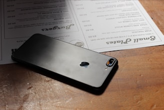 A smartphone with a glass back and an Apple logo rests on a wooden table. Next to it, there are menus with text in various fonts and sizes.