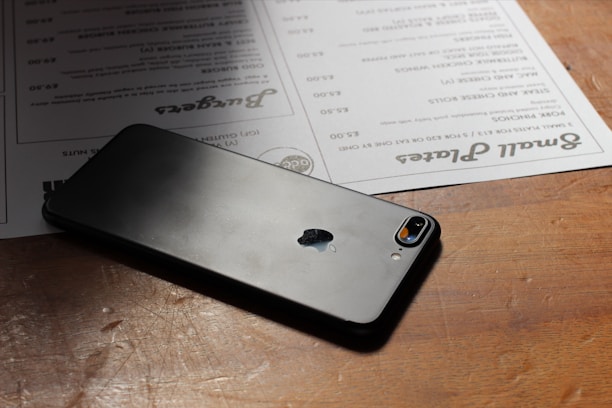 A smartphone with a glass back and an Apple logo rests on a wooden table. Next to it, there are menus with text in various fonts and sizes.