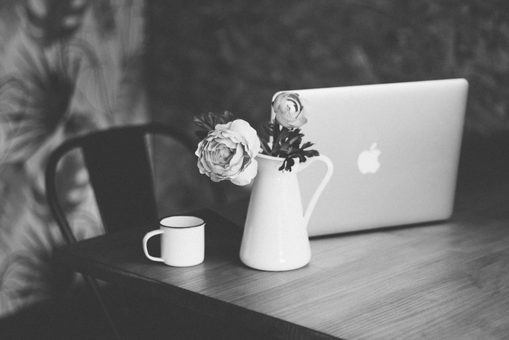 カップの横の花瓶とテーブルの上のMacBookの花びらのセレクティブフォーカスとグレースケー�ル写真