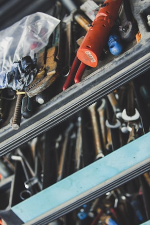 A close-up view of a cluttered toolbox filled with various hand tools, including wrenches, pliers, and a bright red tool. The tools are organized in metal drawers, and the scene has an industrial feel with visible metallic textures and a plastic bag containing additional items.