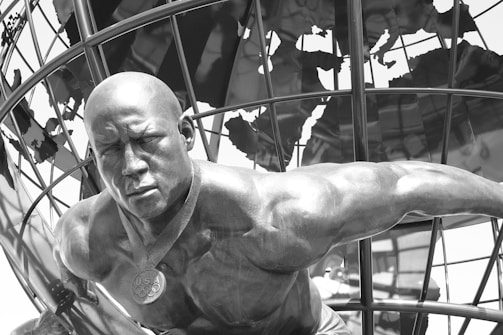 A muscular statue of an athletic figure is prominently positioned in the foreground, wearing a medal with 'USA' inscribed on it. The figure is portrayed leaning forward with a determined expression. In the background, a metallic globe structure is visible, adding an international theme to the setting.