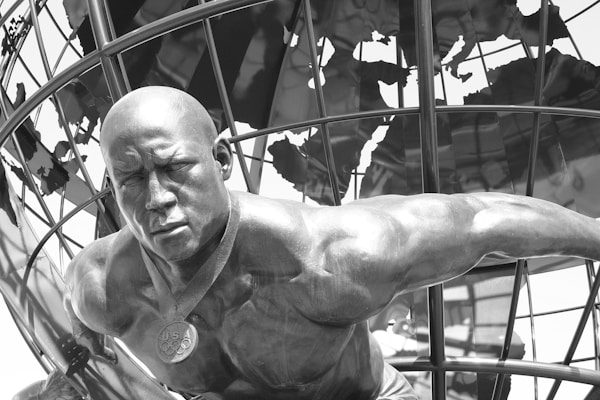 A muscular statue of an athletic figure is prominently positioned in the foreground, wearing a medal with 'USA' inscribed on it. The figure is portrayed leaning forward with a determined expression. In the background, a metallic globe structure is visible, adding an international theme to the setting.