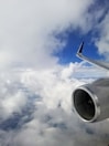 An airplane engine and wing are visible amidst a backdrop of fluffy white clouds and a bright blue sky, suggesting high altitude travel.