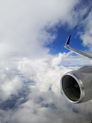An airplane engine and wing are visible amidst a backdrop of fluffy white clouds and a bright blue sky, suggesting high altitude travel.