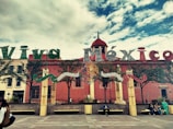 Large decorative letters spell out 'Viva México' in front of a historical building with red walls. The letters are adorned with festive green, white, and red colors. Below the letters, a group of people are seated on benches in a plaza lined with small trees. The sky is partly cloudy, creating a vibrant and lively atmosphere.
