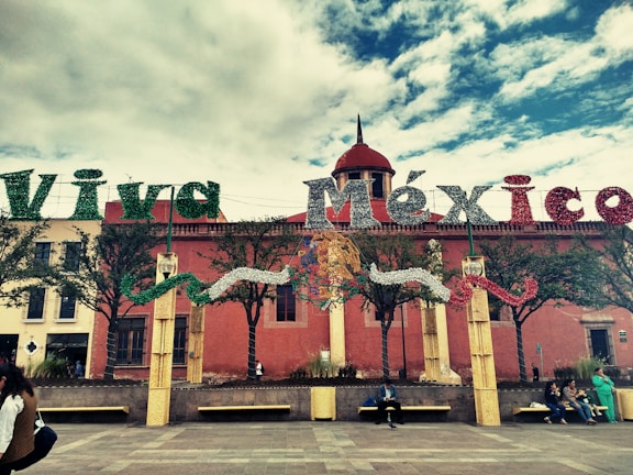 Large decorative letters spell out 'Viva México' in front of a historical building with red walls. The letters are adorned with festive green, white, and red colors. Below the letters, a group of people are seated on benches in a plaza lined with small trees. The sky is partly cloudy, creating a vibrant and lively atmosphere.