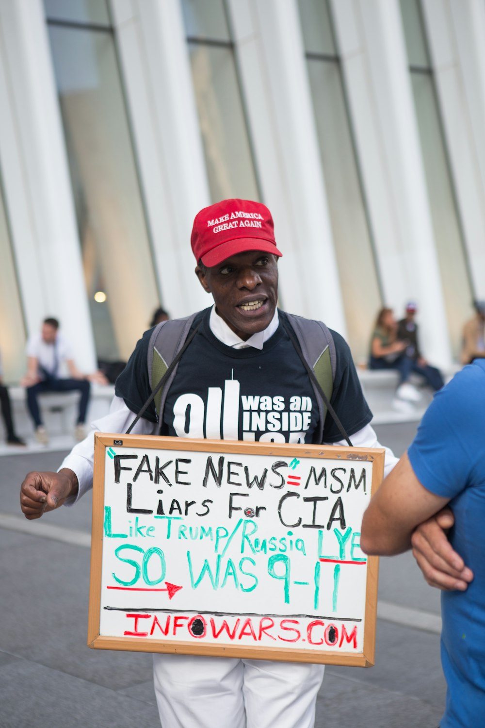 man in black and white shirt with red make America great again cap holding fake news=MSM sign near white building