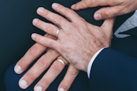 Several hands are placed together, displaying wedding rings on their fingers. The hands appear to be resting on a dark fabric surface and are wearing formal attire.