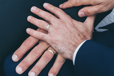 Several hands are placed together, displaying wedding rings on their fingers. The hands appear to be resting on a dark fabric surface and are wearing formal attire.
