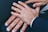 Several hands are placed together, displaying wedding rings on their fingers. The hands appear to be resting on a dark fabric surface and are wearing formal attire.